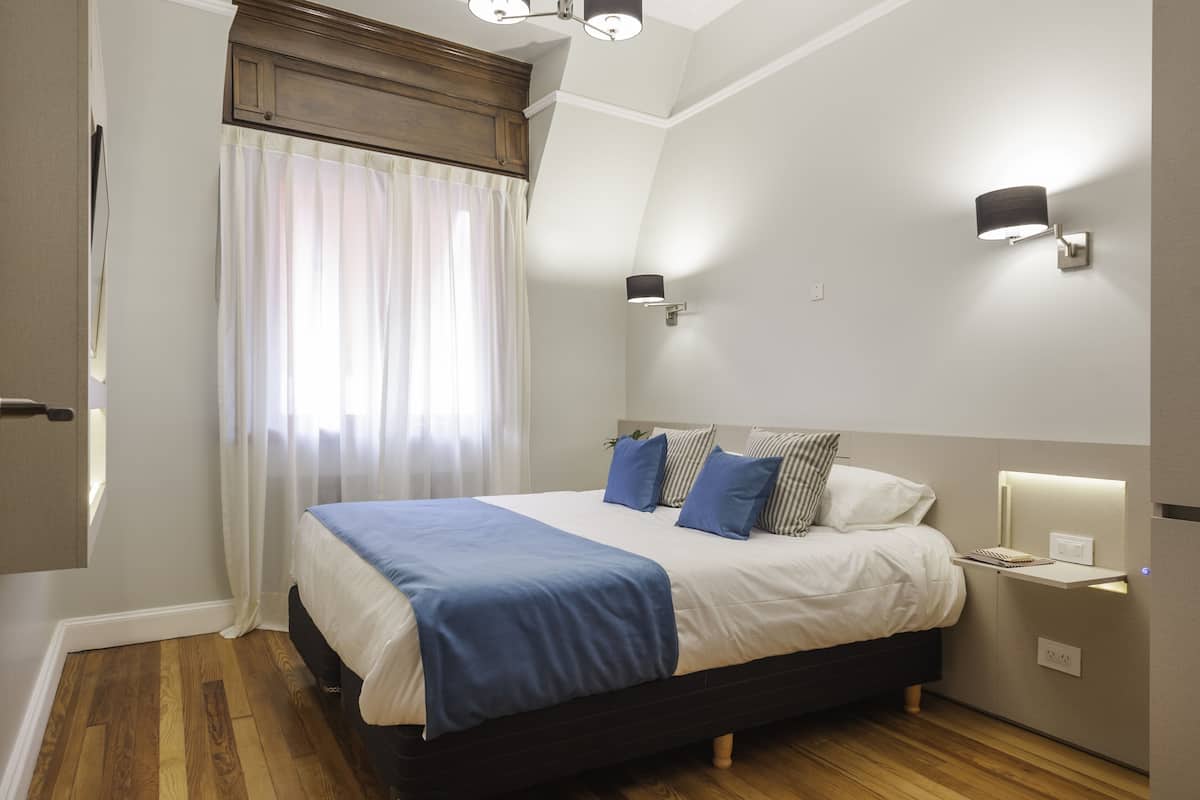 A bedroom with a bed, bedside tables, a nightstand, a window with curtains, and wall-mounted lighting.