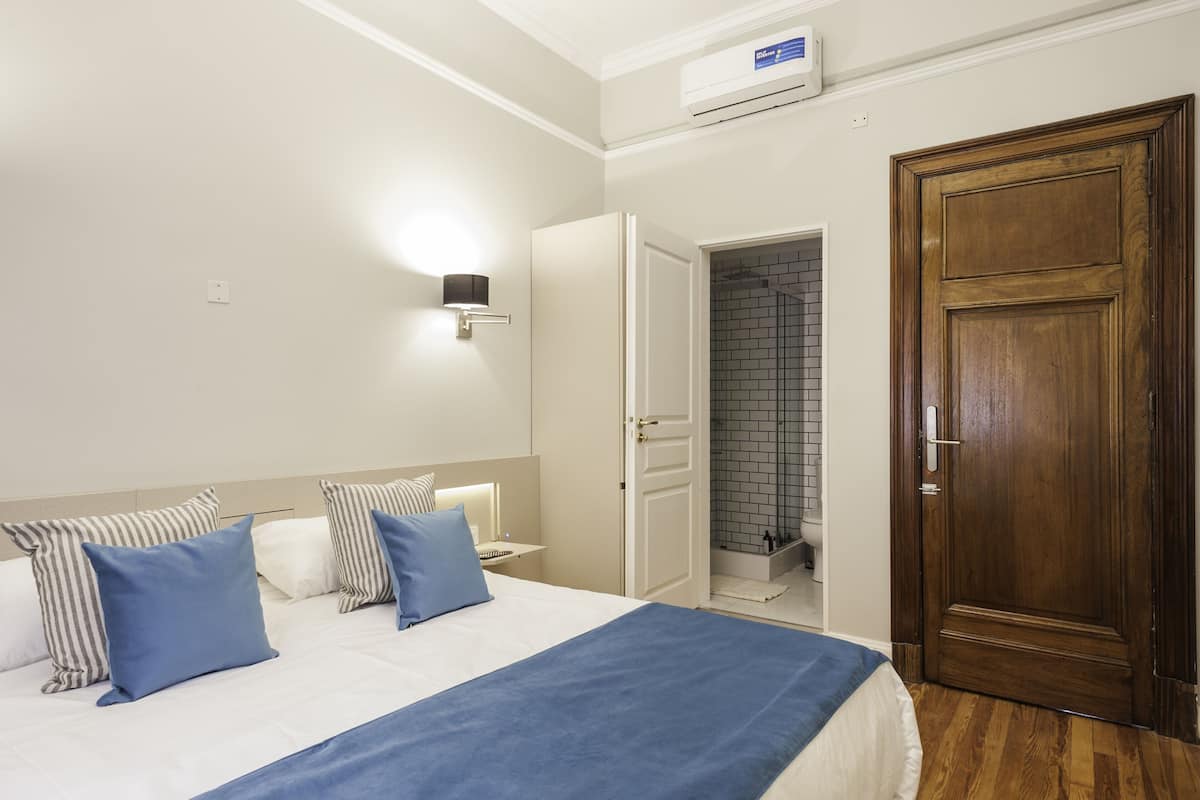 A hotel room with a bed, a wooden door, a bathroom with a tiled shower, and a wall-mounted air conditioner.