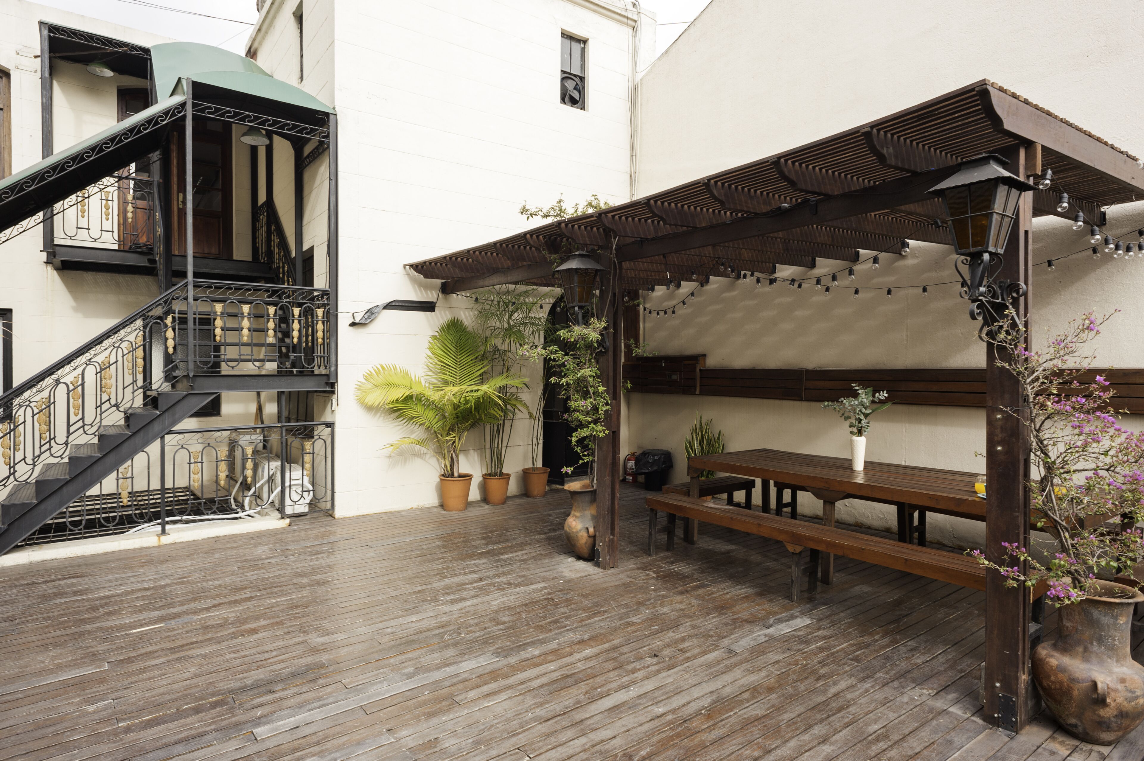 A courtyard with wooden flooring, a wooden bench, potted plants, a pergola with string lights, and a staircase with a metal railing.
