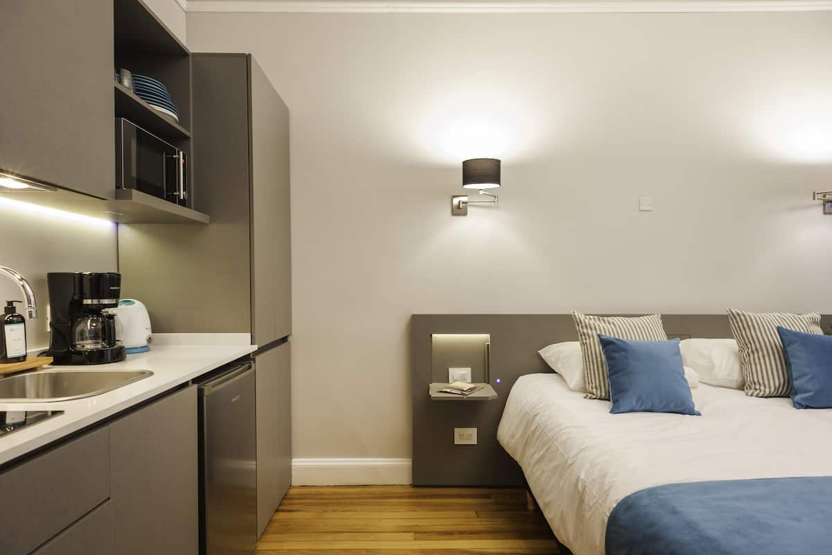 A modern hotel room with a kitchenette, a bed with striped pillows, and a bedside table with a lamp.