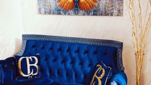 A blue velvet sofa with 'B' cushions, a large artwork of golden wings, and a vase with tall stems. - BALPETEK HOTEL (Istanbul)