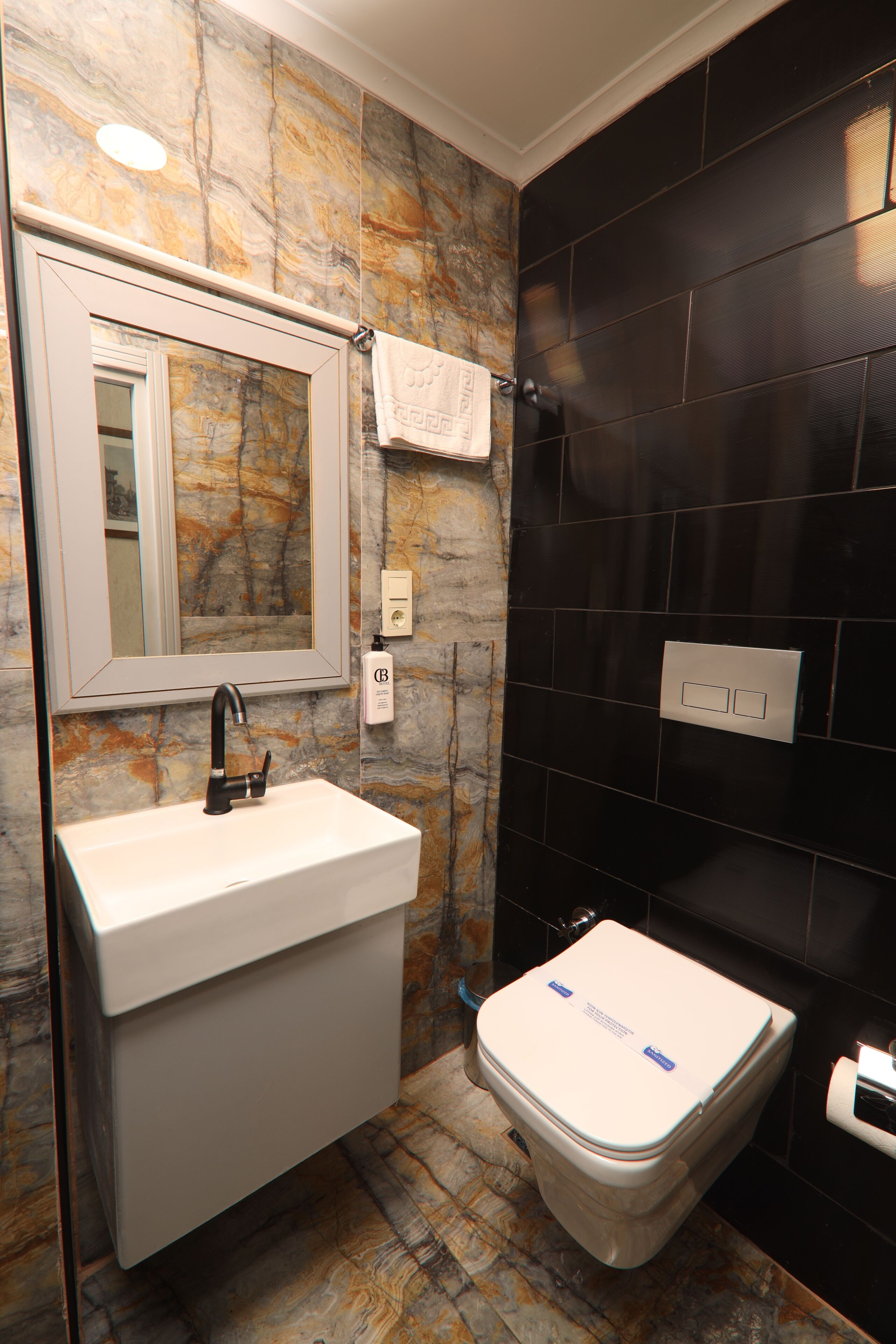 A bathroom with a marble wall, a white sink, a mirror, a towel, and a toilet.