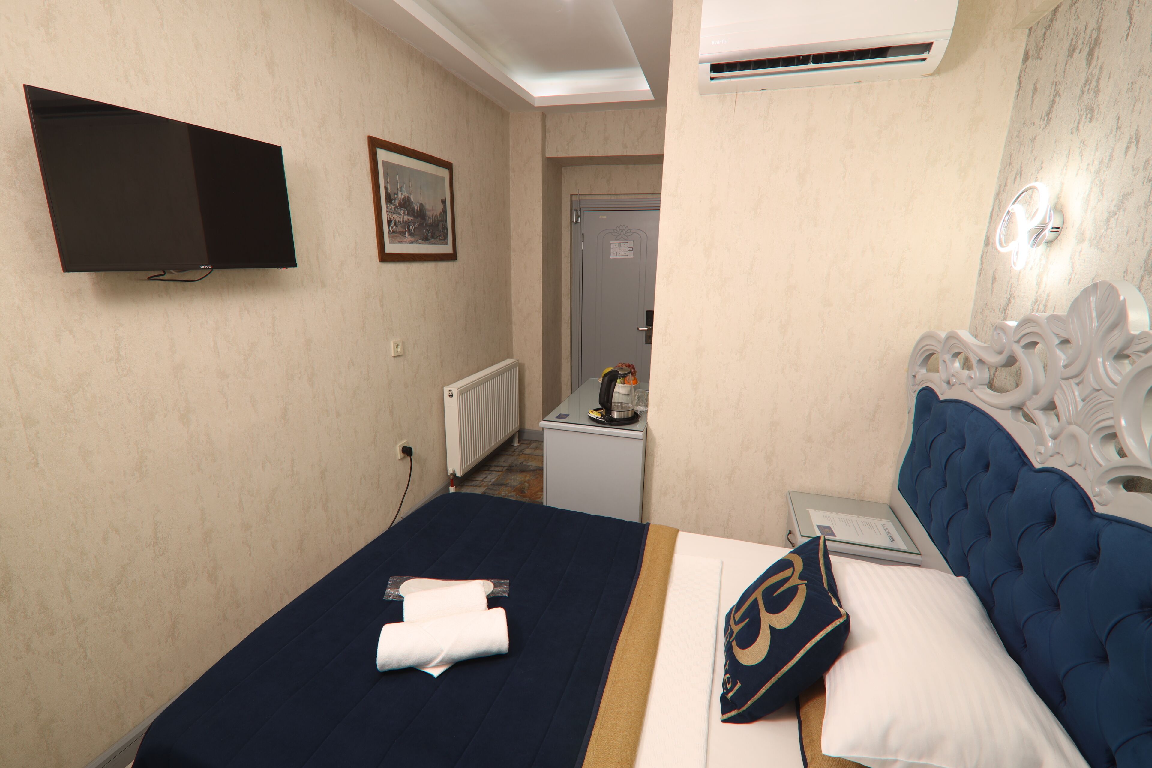 A hotel room with a bed, a flat-screen TV mounted on the wall, a small table with a coffee maker, and a door leading to a bathroom.