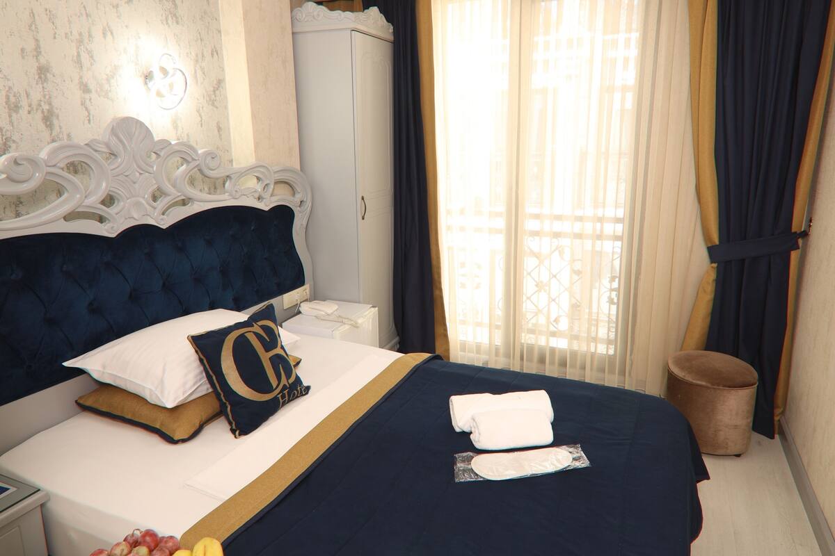 A hotel room with a bed, a bedside table with fruit, a nightstand, a wardrobe, and a window with curtains.