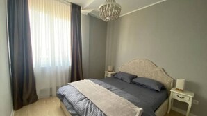 Un dormitorio con cama, mesitas de noche, una araña y una ventana con cortinas.