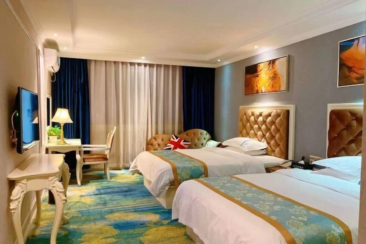 Quarto de hotel com duas camas, uma escrivaninha, uma cadeira, uma televisão e uma janela grande com cortinas.