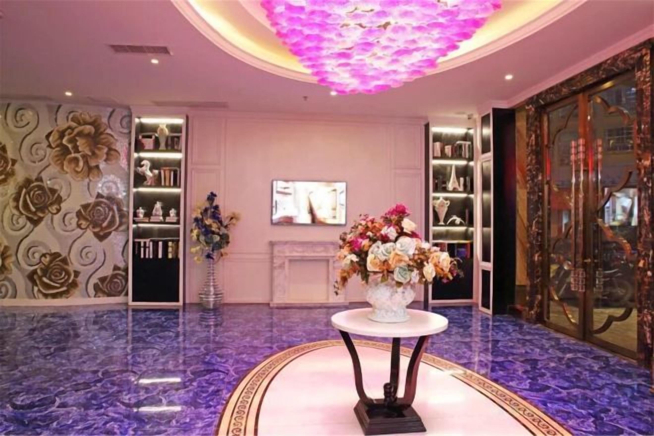 A lobby with a marble floor, a central table with a floral arrangement, and a television screen on the wall.