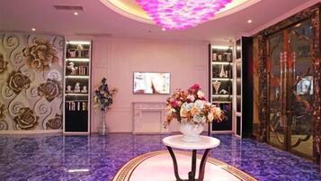 A lobby with a marble floor, a central table with a floral arrangement, and a television screen on the wall.