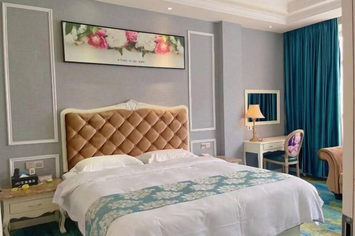 Quarto de hotel com cabeceira estofada, cama com roupa de cama floral, escrivaninha com luminária, uma cadeira e um espelho grande.