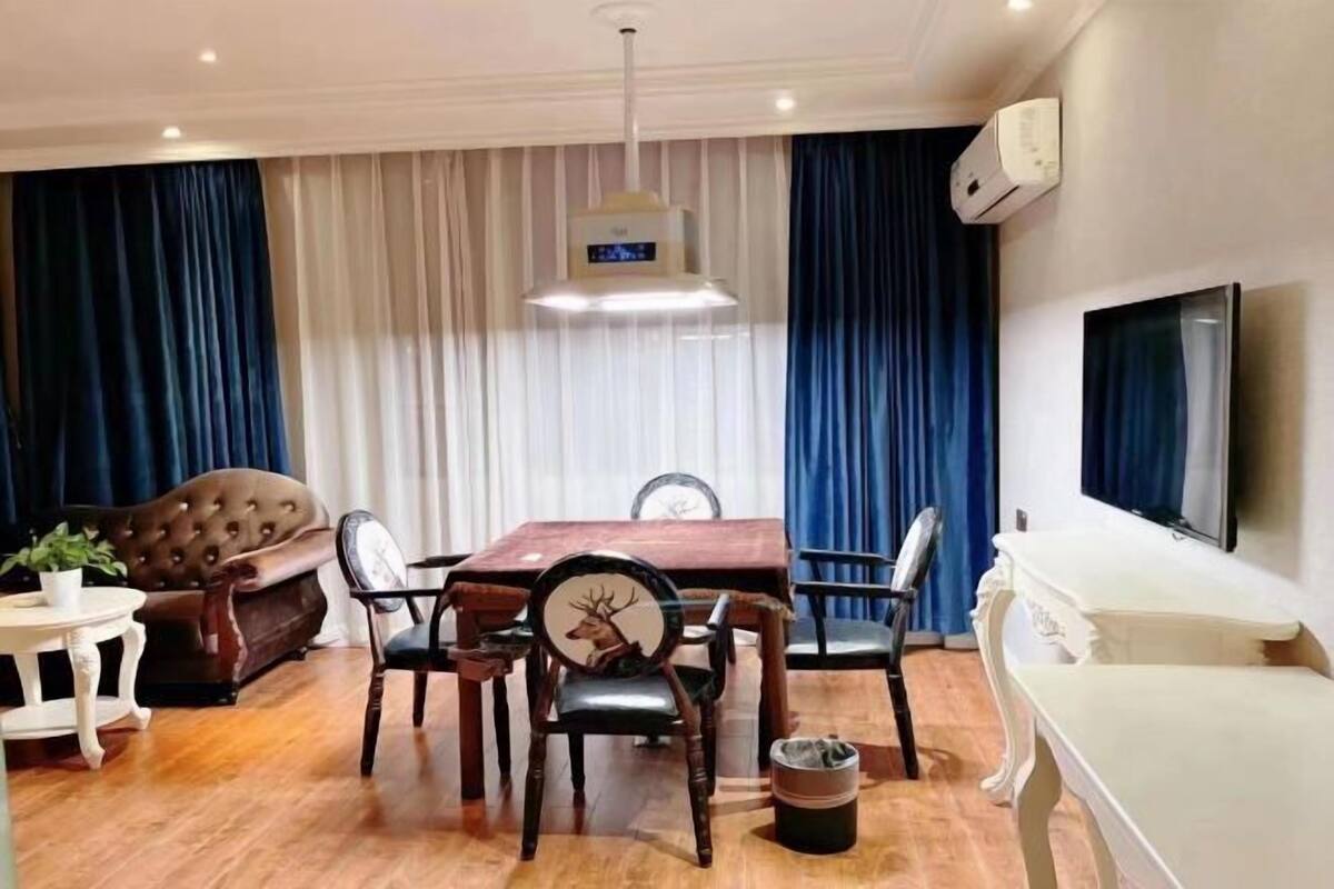 Uma sala de jantar com mesa e cadeiras de madeira, um sofá marrom, uma poltrona branca e uma televisão na parede.
