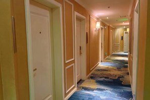Un couloir d’hôtel avec des portes sur le côté gauche, un tapis aux motifs bleus et dorés et un luminaire au plafond.