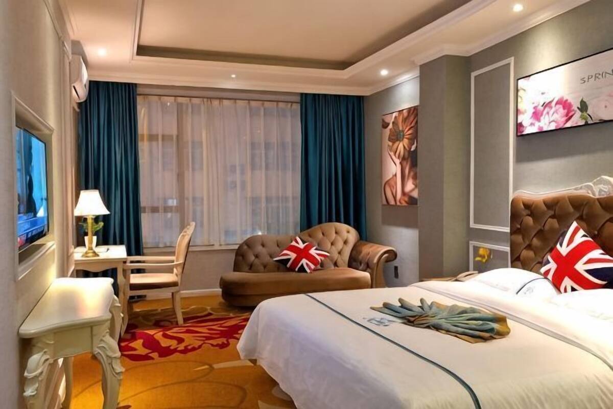 Quarto de hotel com cama, poltrona, escrivaninha, televisão e um quadro na parede.