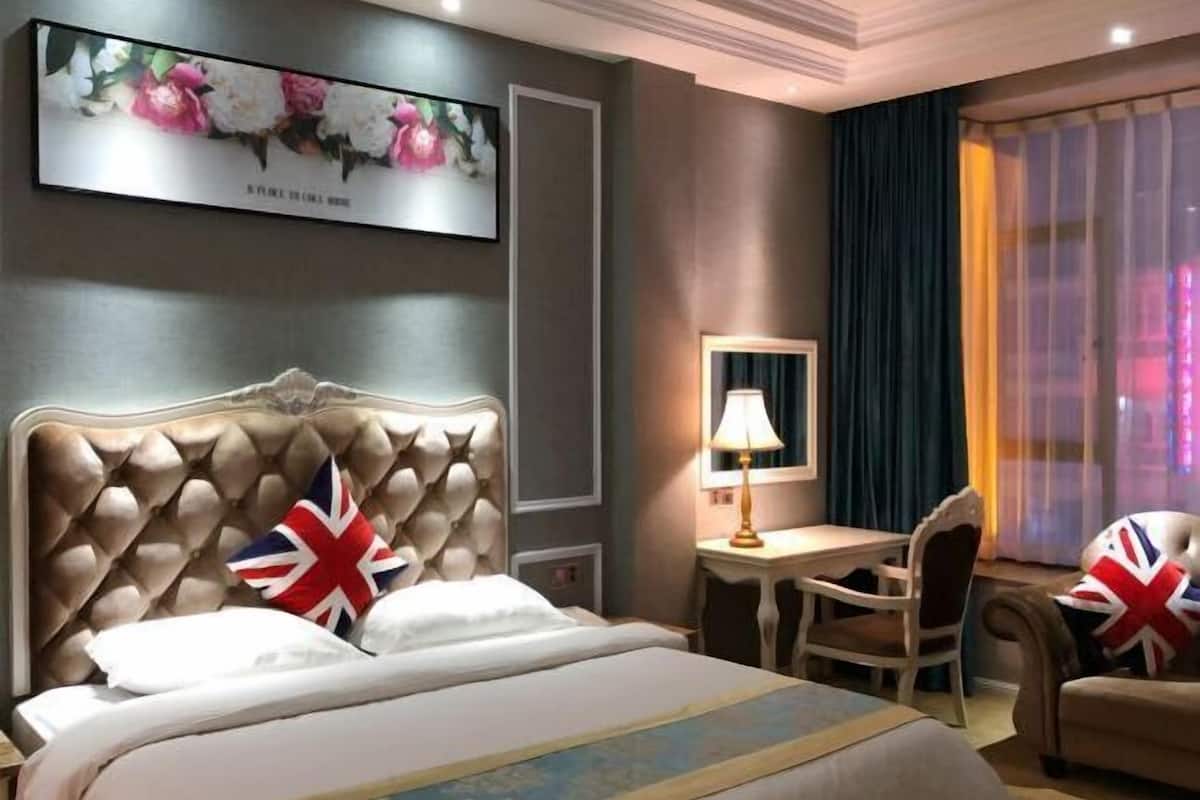 A hotel room with a large bed, a desk, and a chair. There is a painting on the wall and a window with curtains.