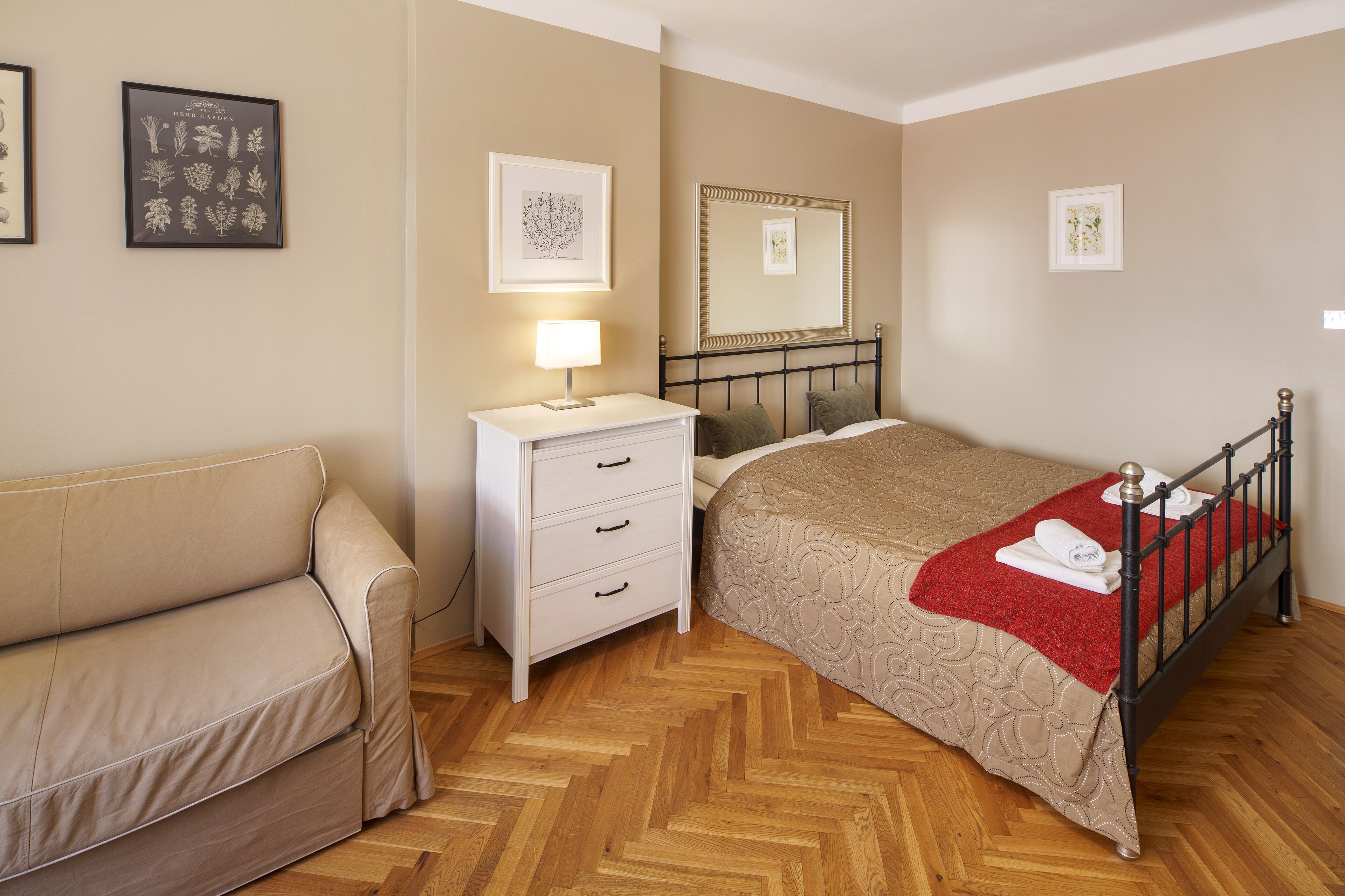 Ein Schlafzimmer mit einem Bett, einem Nachttisch, einer Lampe, einem Spiegel und einem gerahmten Pflanzenmotiv an der Wand.