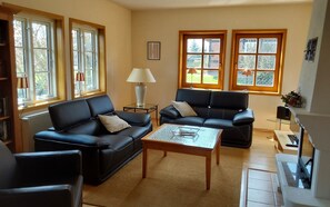A living room with two black leather sofas, a coffee table, and a lamp. There are windows with curtains and a radiator. - Romantische Reetgedeckte Bauernhaushälfte in Alt Rehse (Penzlin OT Alt Rehse)