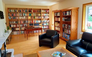 Amplia sala de estar con piso de madera, un sofá de cuero negro, una mesa de centro de madera con revistas, una pequeña mesa con una lámpara y una estantería llena de libros.