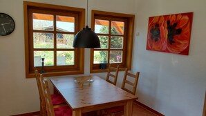 Zona pranzo con tavolo e sedie in legno, due finestre con vista sul verde, una lampada pendente nera e un quadro floreale rosso al muro.