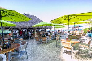 An outdoor café with thatched-roof tables and green umbrellas, surrounded by chairs and a pool area.