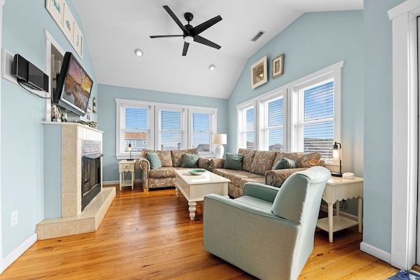 A living room with a fireplace, a ceiling fan, and a large window with blinds.