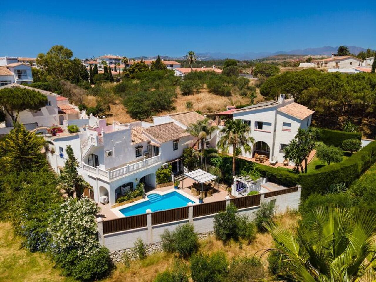 A large house with a swimming pool, surrounded by lush greenery and a clear blue sky.