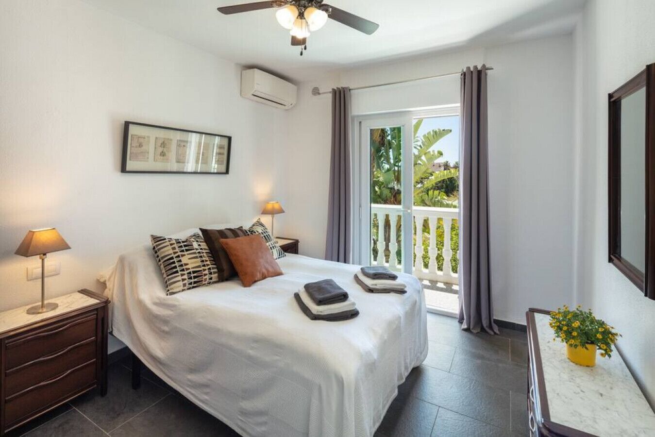 Ein Hotelzimmer mit einem Bett, zwei Nachttischlampen, einem Deckenventilator, einer Klimaanlage, einem kleinen Tisch mit einer Topfpflanze und einem Balkon mit Vorhängen.