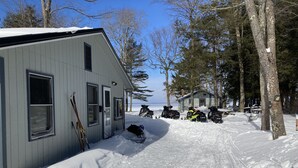 A cabin with a snow-covered driveway, a sled leaning against the wall, and snowmobiles parked outside. - 4 seasons of adventures on Indian Lake/Sunset View Resort/Unit#4 (Manistique)