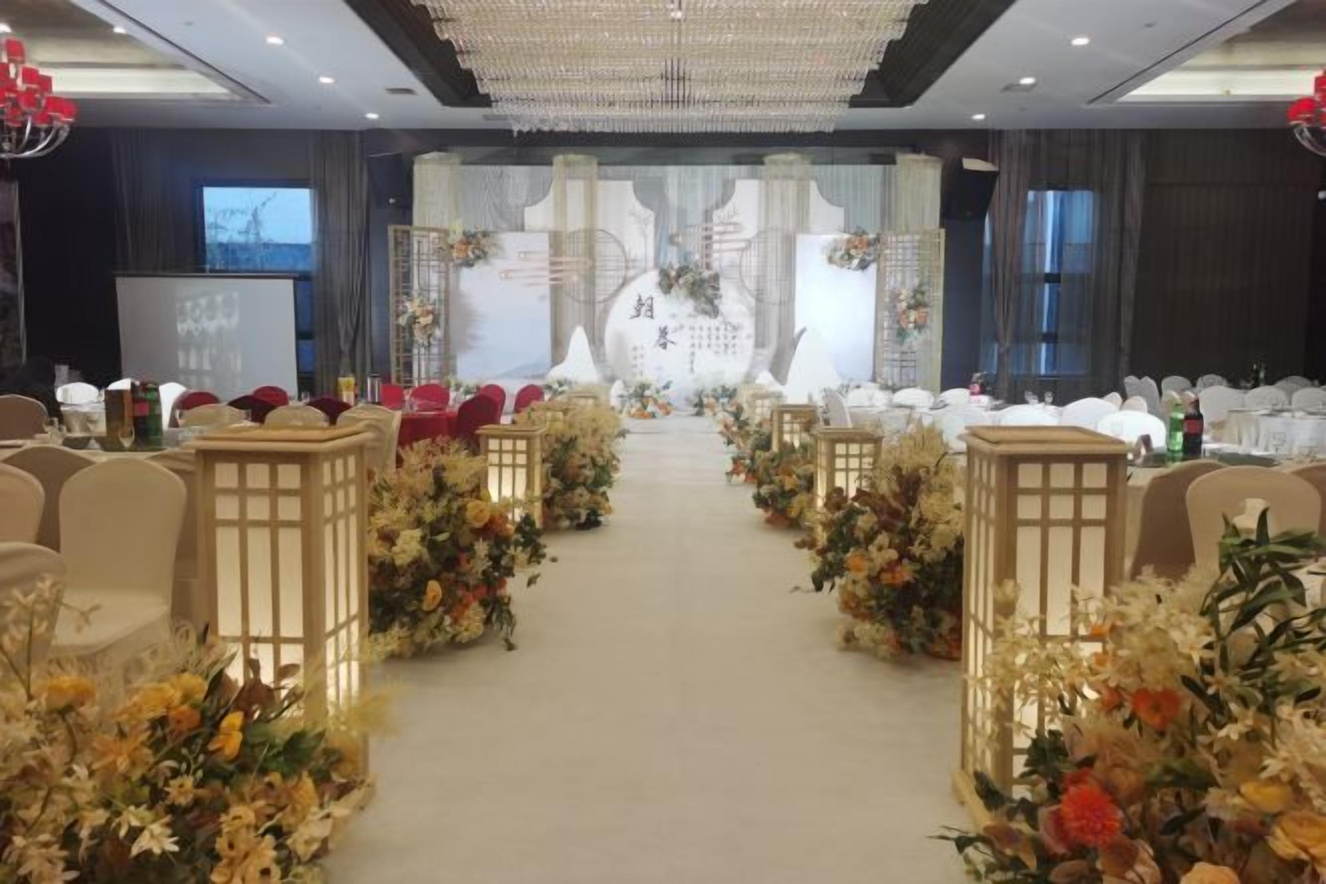A banquet hall with a central aisle flanked by tables, a stage with floral decorations, and a chandelier.