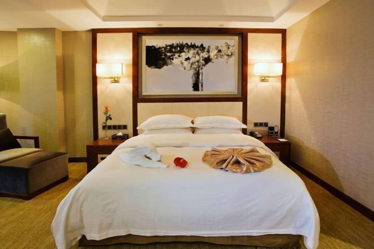 A hotel room with a large bed, two bedside lamps, a painting above the bed, a small table, and a chair.