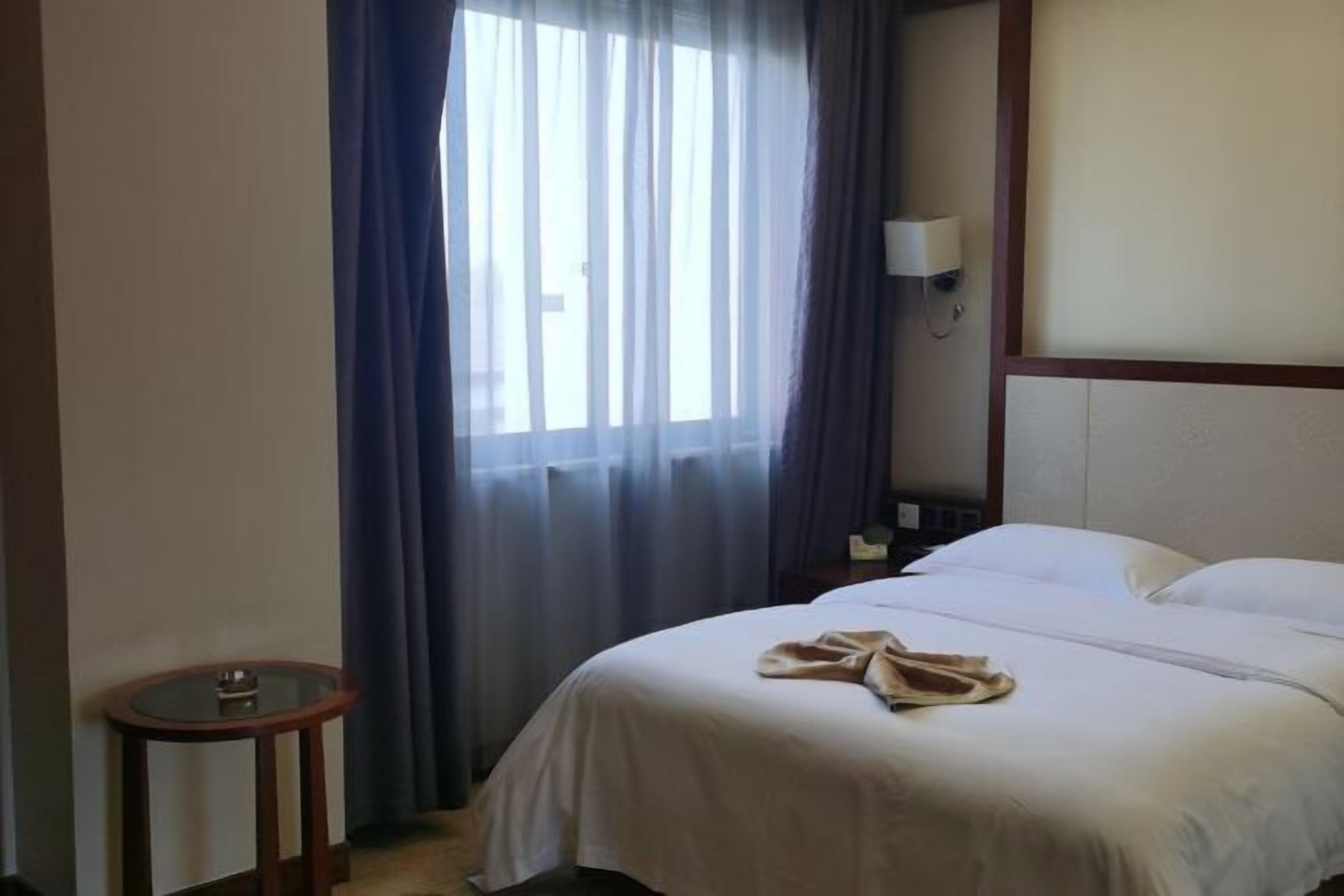 Hotellihuone, jossa on sänky, yöpöytä, lamppu ja ikkuna, jossa on verhot.