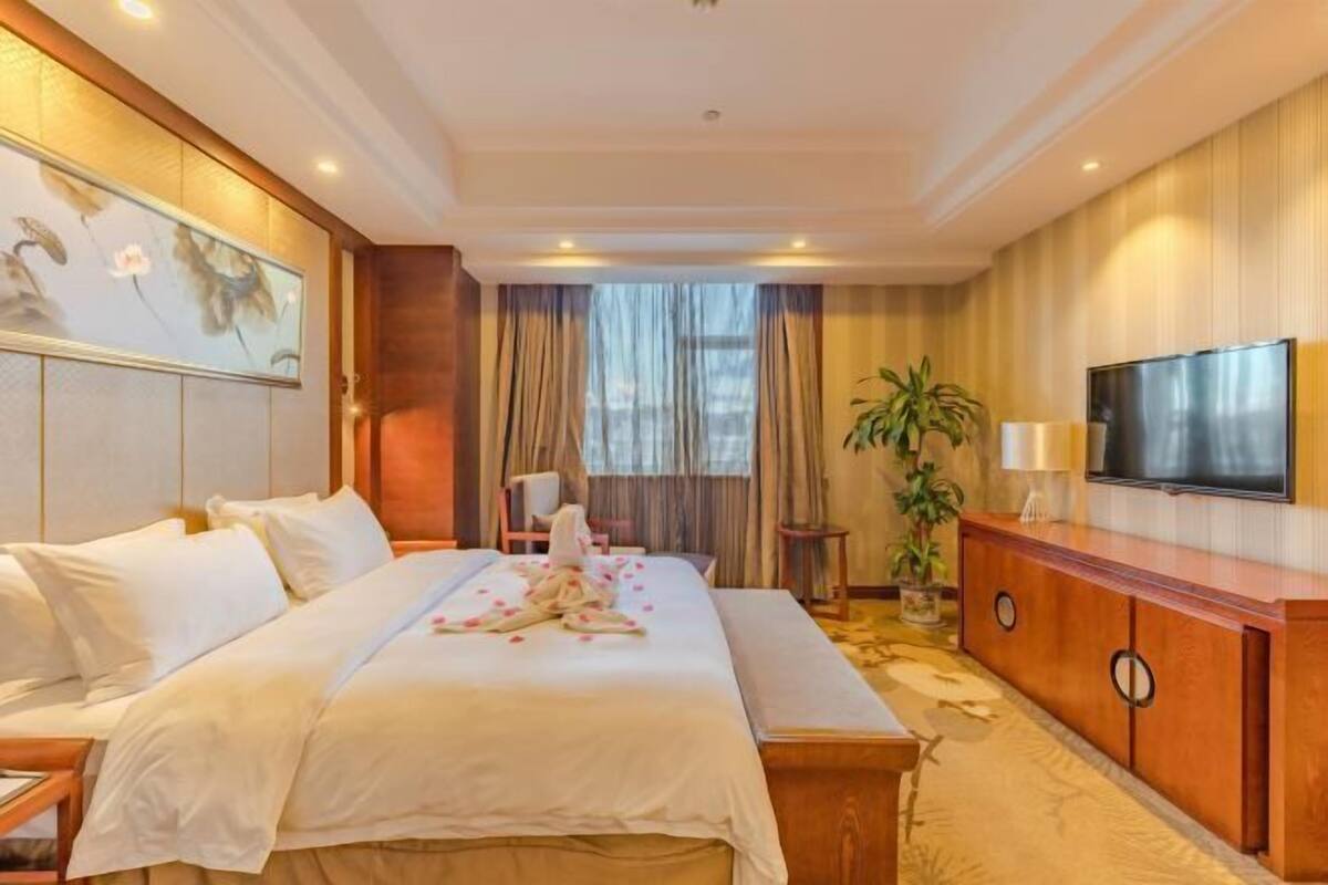 A hotel room with a bed, a TV, a wooden cabinet, and a large window with curtains.