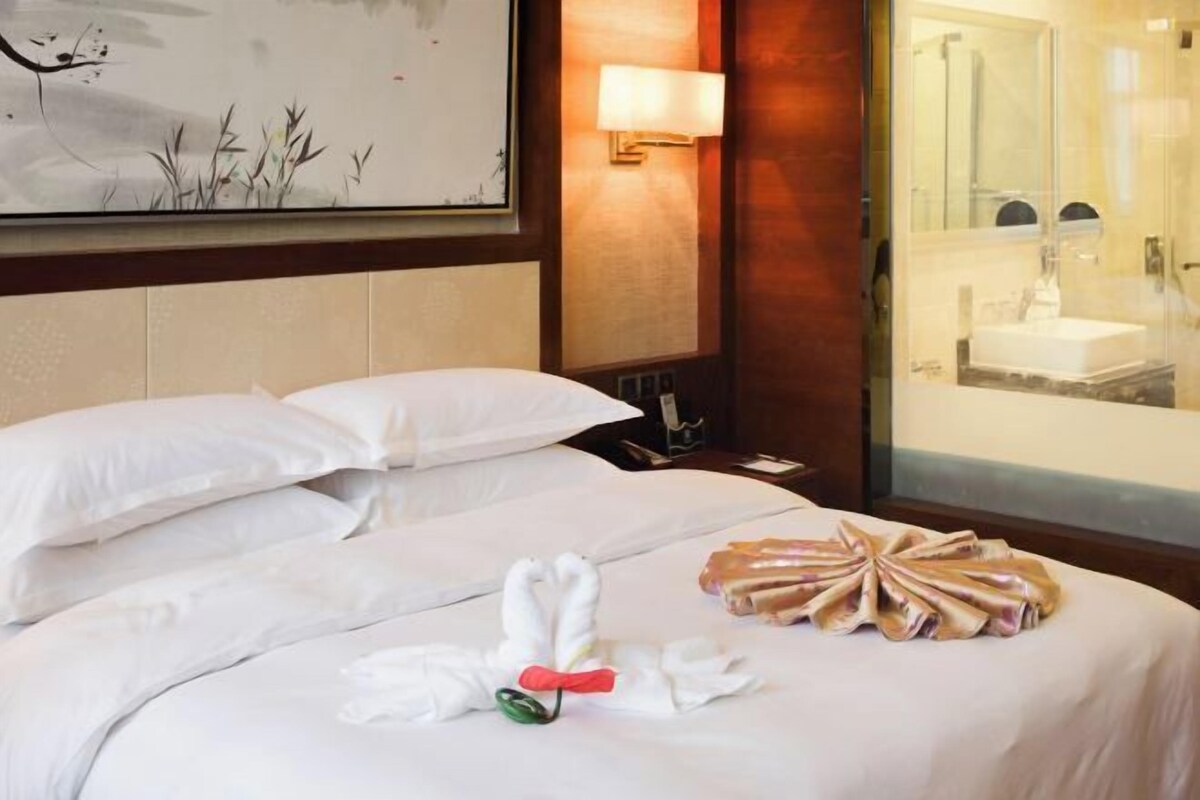 Une chambre d’hôtel avec un lit soigneusement fait, une lampe fixée au mur et une salle de bain visible à travers une porte en verre.