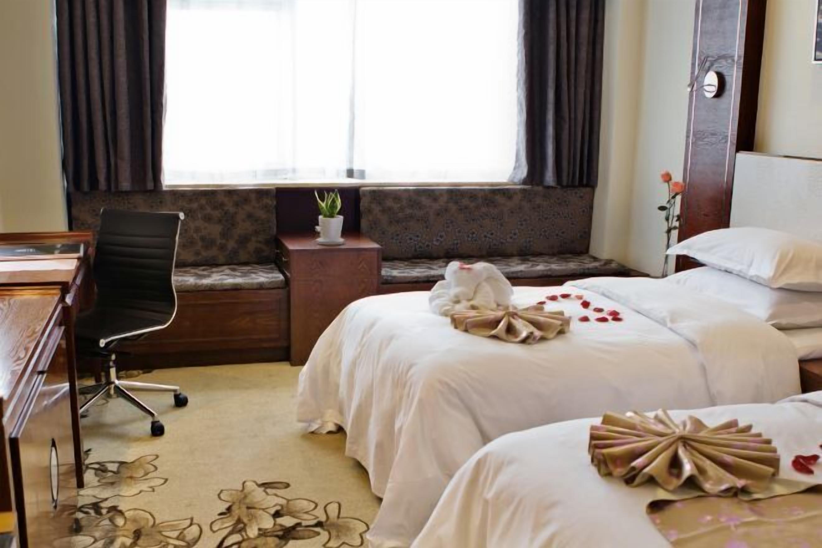 Habitación de hotel con dos camas, un escritorio, una silla, una mesita con una planta y una ventana con cortinas.