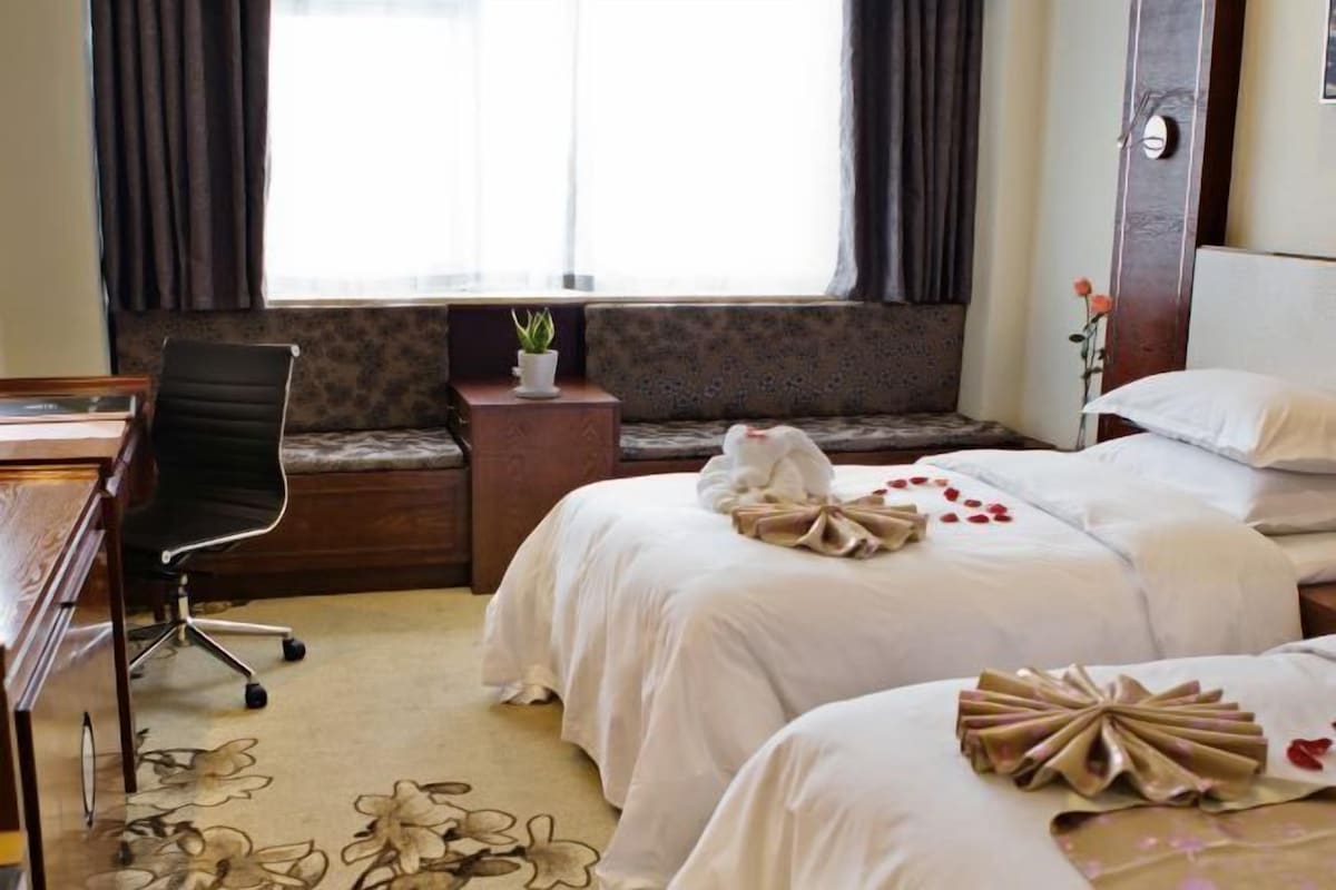 Une chambre d’hôtel avec deux lits, un bureau, une chaise, une petite table avec une plante et une fenêtre avec des rideaux.