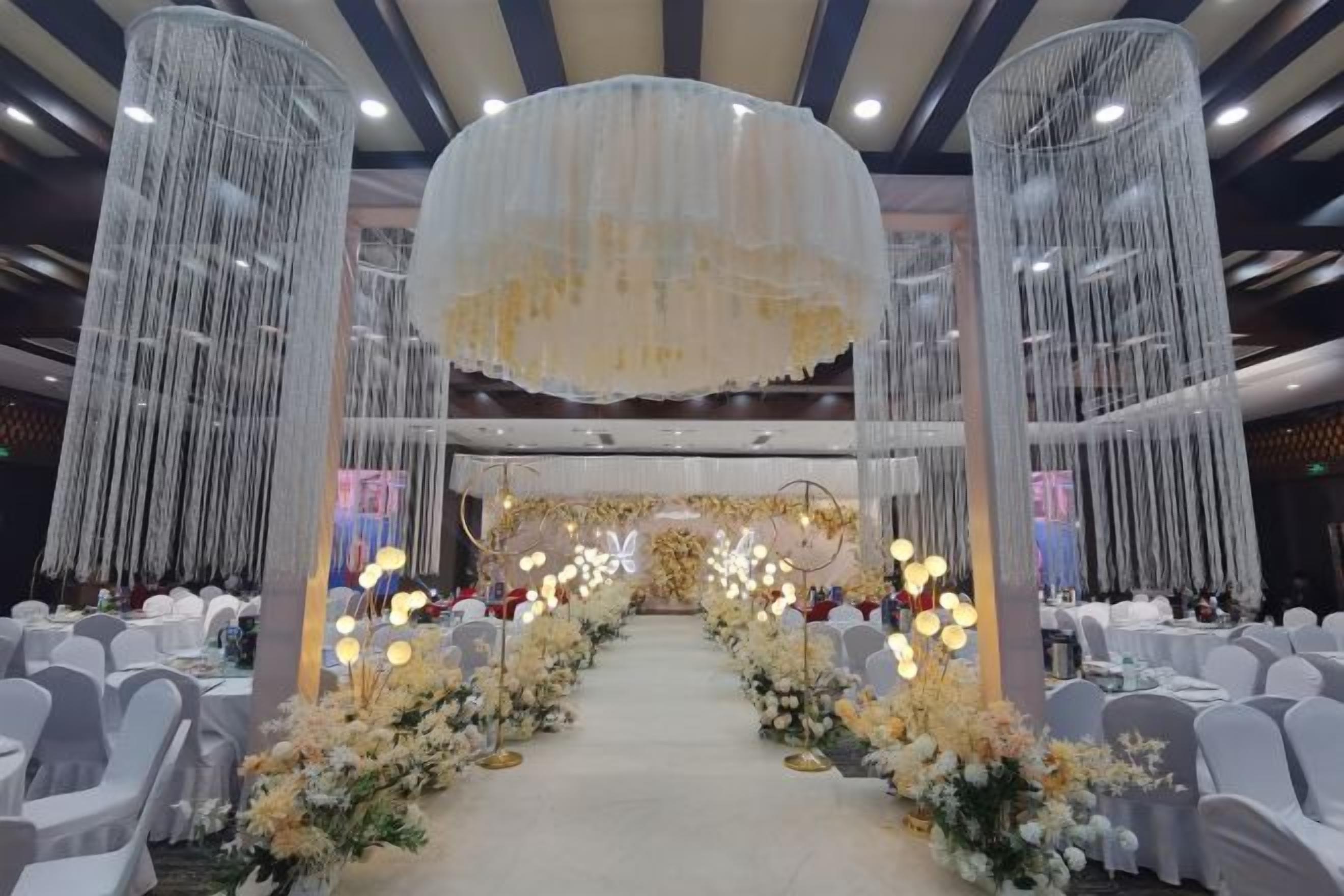 A banquet hall with a central aisle, white chairs on both sides, and a floral arrangement along the aisle.