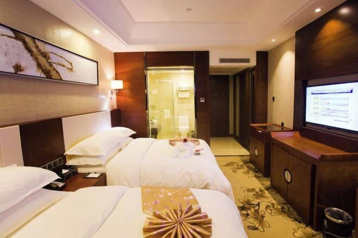 A hotel room with two beds, a TV, a desk with a computer, and a bathroom visible through a glass door.