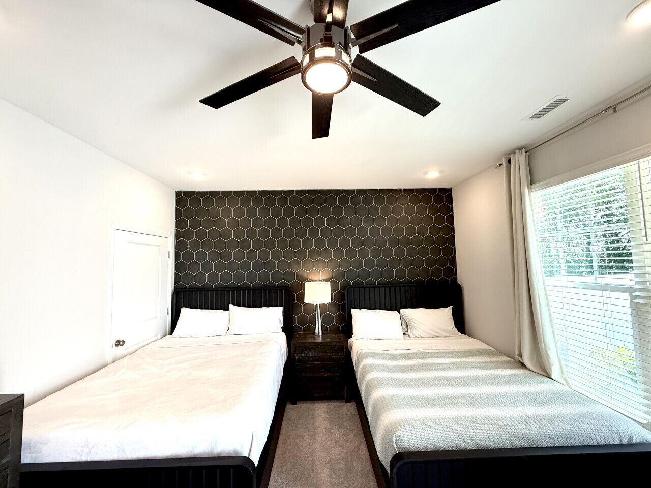 Une chambre d’hôtel avec deux lits, un ventilateur de plafond, une lampe fixée au mur et une fenêtre avec des stores.