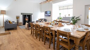 Un comedor amplio con una larga mesa de madera dispuesta para una comida, rodeada de sillas de madera. Se aprecia una chimenea de diseño moderno al fondo, y un televisor está montado en la pared.