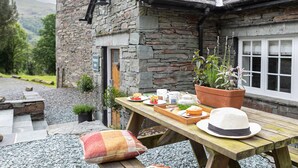 Un edificio de piedra con puerta y ventana de madera, una mesa de picnic con comida y un sombrero, y un jardín con plantas.