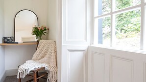 Un cuarto acogedor con una silla de madera cubierta con una manta blanca de punto, una mesita con un libro y un objeto decorativo, un espejo y una ventana con vistas a la vegetación.