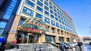 A multi-story hotel with a glass facade, a prominent entrance with a curved awning, and a parking lot with cars. - Junrui Garden Hotel (Qianguo)