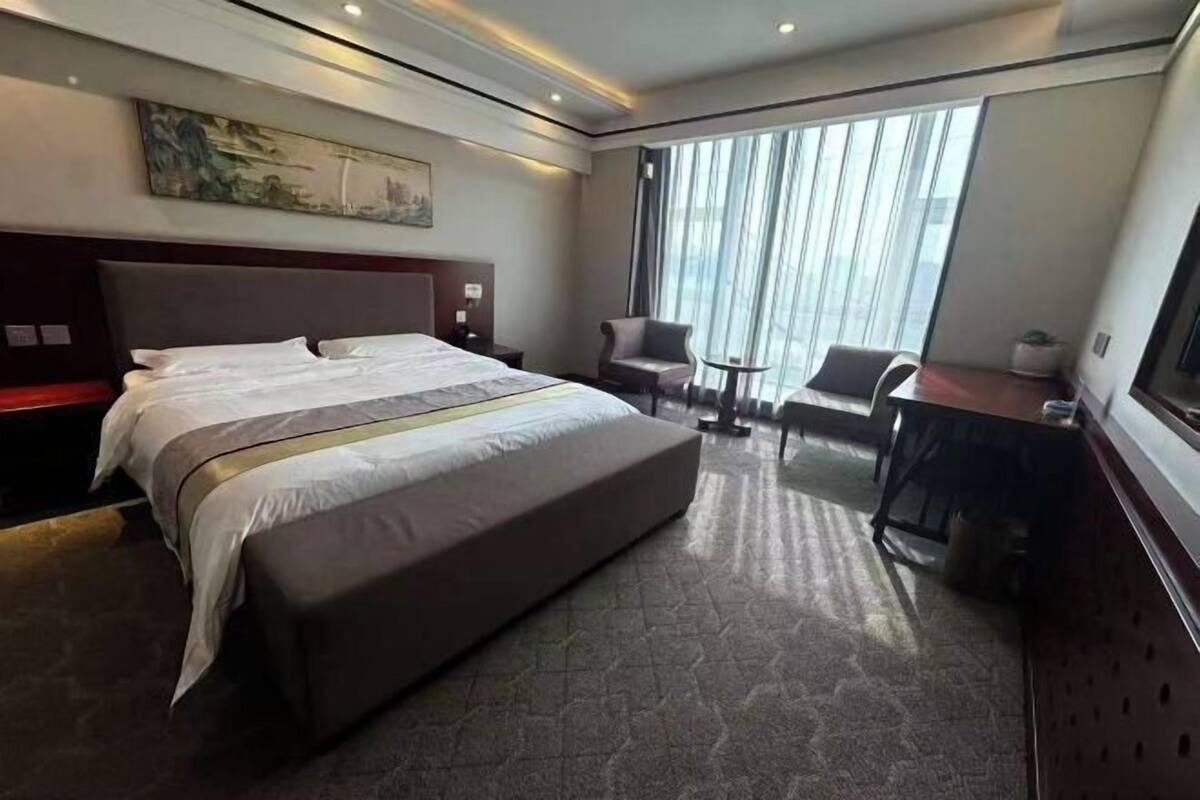 Ein Hotelzimmer mit einem großen Bett, einem Schreibtisch, einem Sessel und Blick auf die Stadt durch das Fenster.