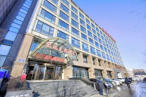 A multi-story building with a large glass facade, a prominent entrance with a curved overhang, and a parking area with cars. - Junrui Garden Hotel (Qianguo)