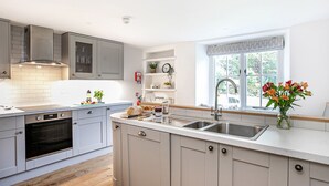 A modern kitchen with grey cabinets, a built-in oven, a large island with a sink, and a window with a view of greenery. - Little How (Ambleside)