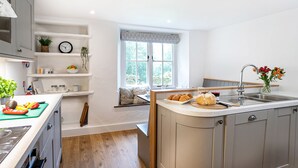 A modern kitchen with white cabinetry, a wooden floor, a built-in bench, and a window with a view of greenery. - Little How (Ambleside)