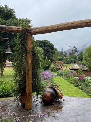 Une pergola en bois dans un jardin, agrémentée d’une cloche, de plantes en pot et offrant une vue sur un quartier résidentiel en arrière-plan.