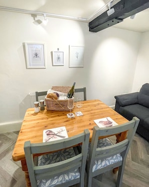 Un comedor con una mesa de madera, sillas, una cesta de pan, saleros y pimenteros, dos copas de vino y tres obras de arte enmarcadas en la pared.