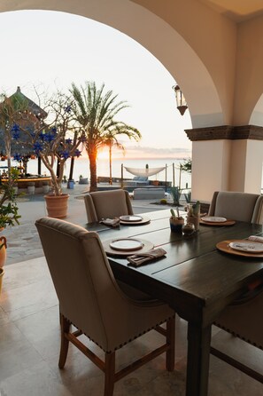 Een terras met een tafel voor vier personen, omringd door stoelen, met uitzicht op zee en palmbomen op de achtergrond.