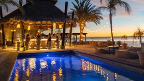 A pool area with a thatched-roof structure, palm trees, and a view of the ocean.