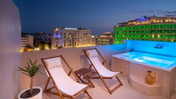 Een terras op het dak met twee lounge stoelen, een kleine tafel en een jacuzzi. Op de achtergrond een stadsgezicht met verlichte gebouwen.