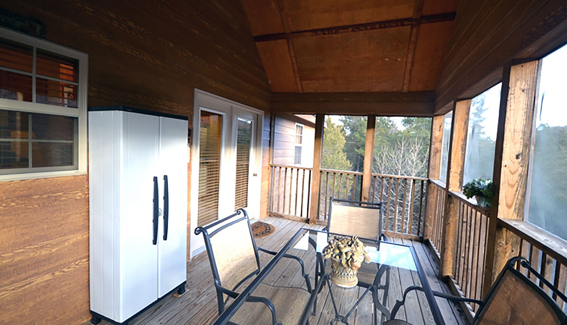 En inglasad veranda med ett trädäck, ett glasbord, metallstolar, ett vitt kylskåp och en dörr som leder ut.